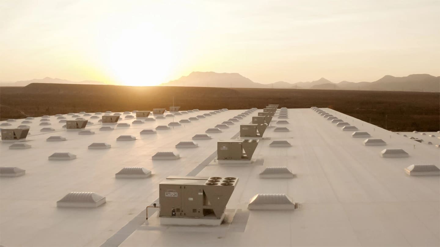 Rows of packaged rooftop units and skylights on a white membrane roof of a large distribution warehouse at sunset, desert mountains on the horizon